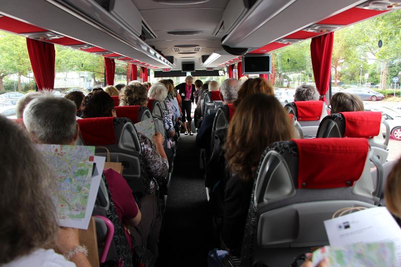 Eine Gruppe an Senioren unternimmt eine Busfahrt, die Stadtführerin steht vorne im Bus und erzählt.